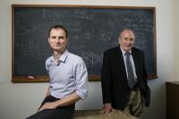 Two professors sit in front of a blackboard.